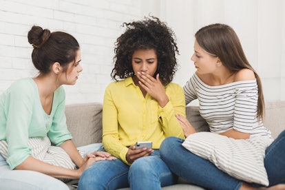 Shocked girl looking at phone, friends calming her sitting on couch during home party