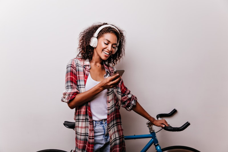 Enchanting stylish woman in headphones reading phone message. Well-dressed african girl with bicycle...