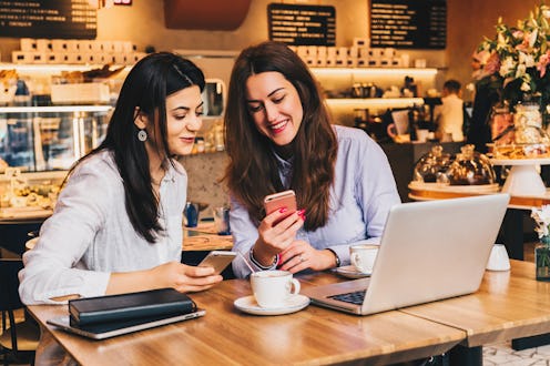 Two young happy women are sitting in cafe at table in front of laptop, using smartphone and laughing...