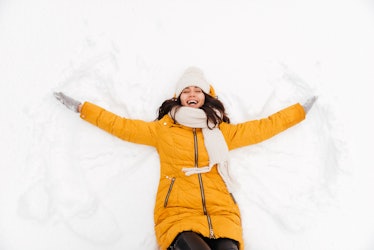 Happy playful brunette lady lying on snow and making snow angel figure with hands and legs