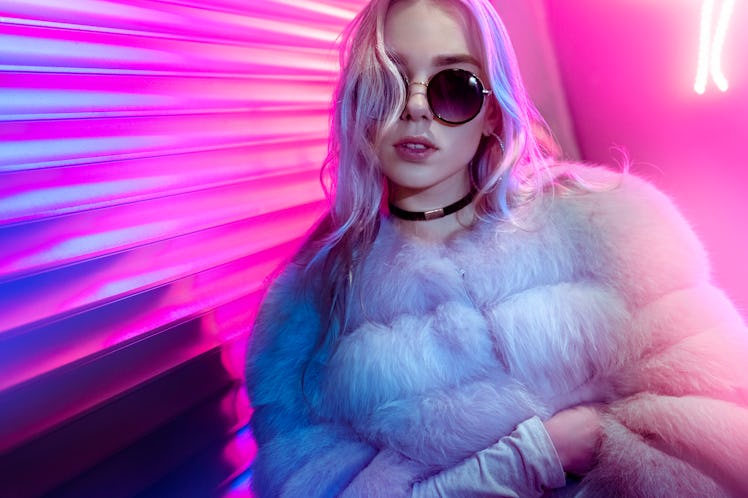 Teen hipster girl in stylish glasses and fur posing on street neon light wall background, female tee...