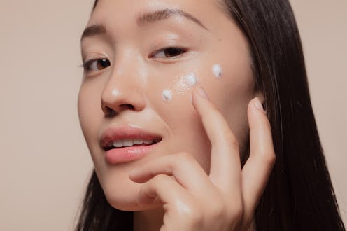 Close up of a young woman applying moisturizer to her face. Asian woman looking happy while followin...
