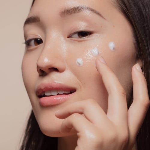 Close up of a young woman applying moisturizer to her face. Asian woman looking happy while followin...