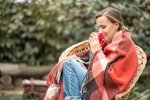 Beautiful girl resting and drinking coffee sitting in autumn garden in chair wrapped in a plaid wool...