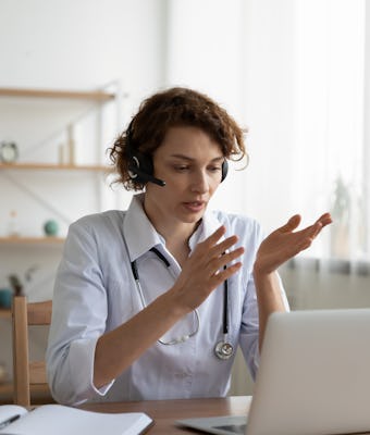 Female general practitioner wears white coat and headset speaking videoconferencing on laptop comput...