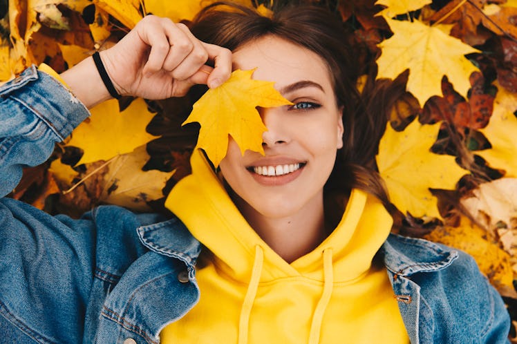 Autumn walk. Woman portrait. Happy girl in yellow hoodie and jean jacket is playing with leaf, looki...
