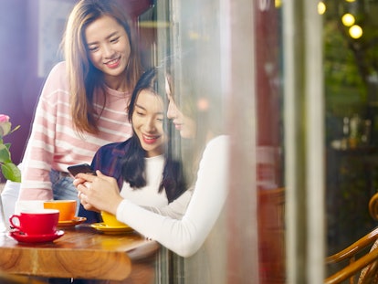 three happy beautiful young asian women sitting at table chatting talking playing with cellphone in ...