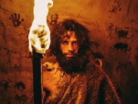 Portrait of Primeval Caveman Wearing Animal Skin Standing in His Cave At Night, Holding Torch with F...