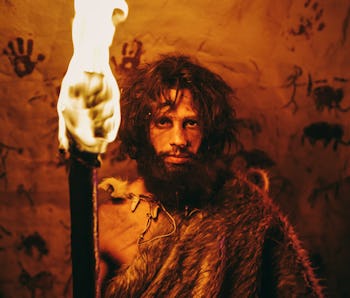Portrait of Primeval Caveman Wearing Animal Skin Standing in His Cave At Night, Holding Torch with Fire. Primitive Neanderthal Hunter / Homo Sapiens At Night Alone. In the Background Cave Art Drawings
