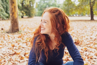 Cute happy young woman with tousled red hair relaxing in a park in autumn sitting amongst the fallen...