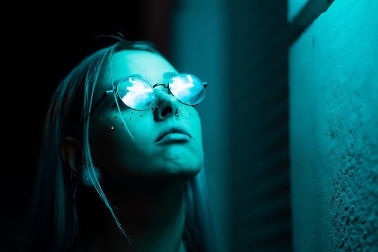 Millennial enigmatic pretty girl with unusual dyed hairstyle near glowing neon wall at night. Blue h...