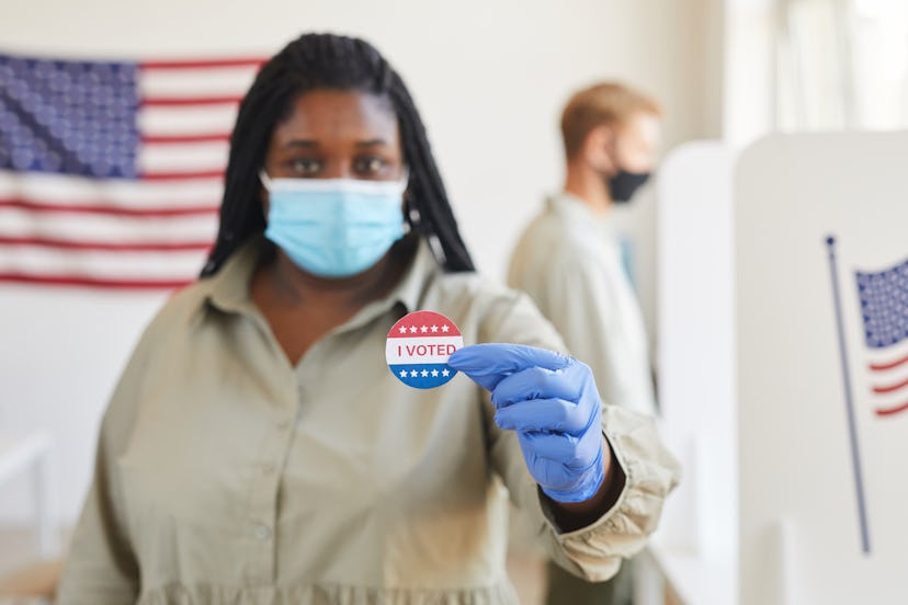 Blurred portrait of African-American woman holding I VOTED sticker while standing t polling station ...