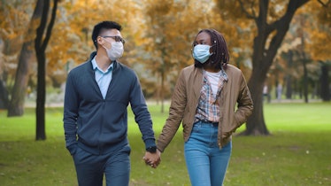 Multiethnic couple in protective masks holding hands walking in park. Lovely diverse man and woman o...