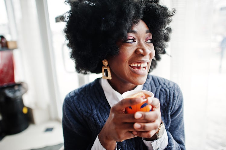 Curly hair african american woman wear on sweater posed at cafe indoor with cup of tea or coffee.