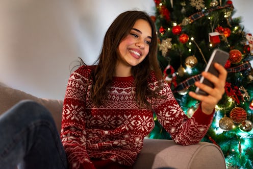 Woman texting on smartphone with christmas tree. Happy woman reading text on a phone on christmas ly...