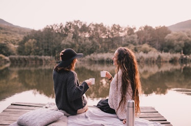 Two female friends in knitted warm sweaters having picnic near lake with autumn forest and lake on t...