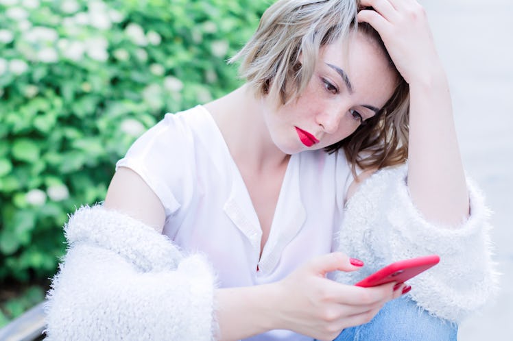 Attractive girl with red lips and manicure is sitting in the park drinking coffee and holding red ph...