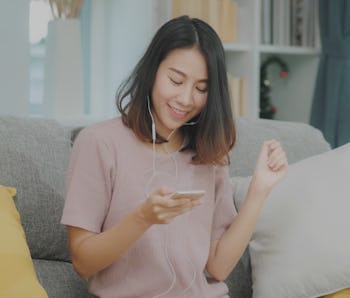 Asian woman listening music and using smart phone, female using relax time lying on home sofa in liv...