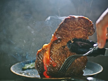Cooking pan seared ribeye steak in nonstick teflon coating pan with kitchen tongs and fork on dark background with smoke and fog. People enjoy home cooking lifestyle concept /close up, selective focus