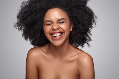 Happy young African American woman with bare shoulders laughing against gray background while repres...