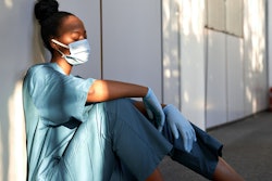 Tired exhausted female african scrub nurse wears face mask blue uniform gloves sits on hospital floo...