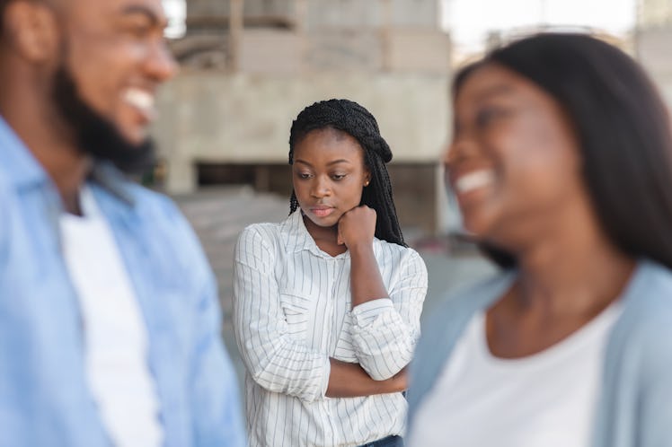 Third wheel. Upset black girl jealous to her dating friends, standing alone on background outdoors, ...