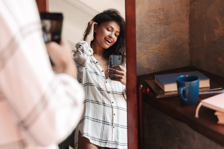 Photo of joyful brunette woman wearing shirt taking selfie photo and looking at mirror in apartment