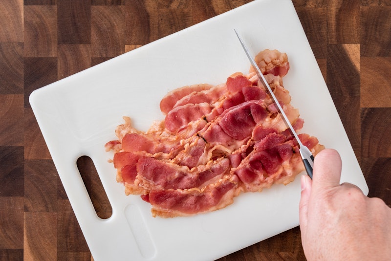 Pre-cooked bacon on white cutting board with woman's hand holding knife cutting bacon, on wood butch...
