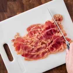 Pre-cooked bacon on white cutting board with woman’s hand holding knife cutting bacon, on wood butch...