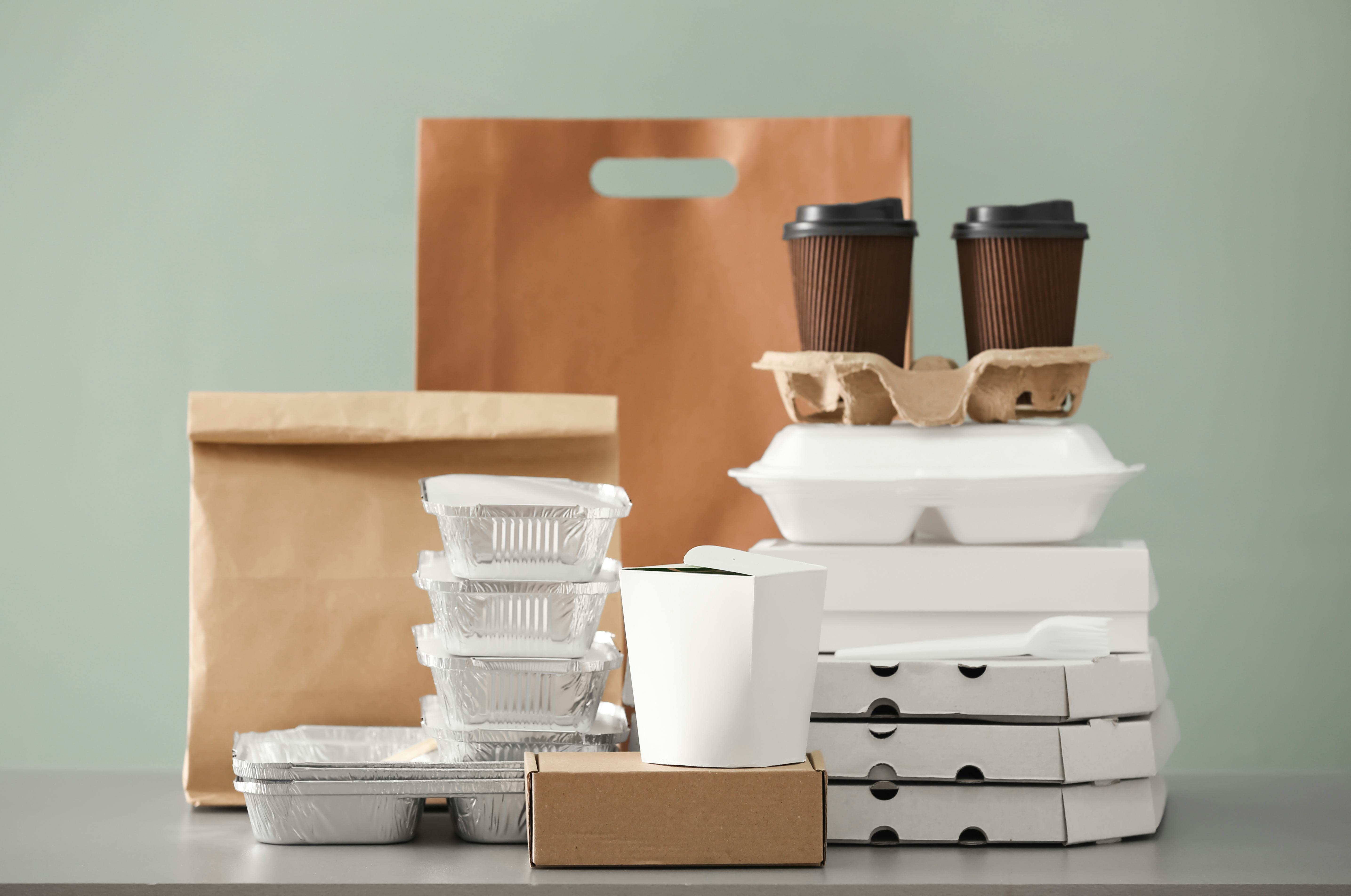 Different packages and carton cups on table against color background. Food delivery service