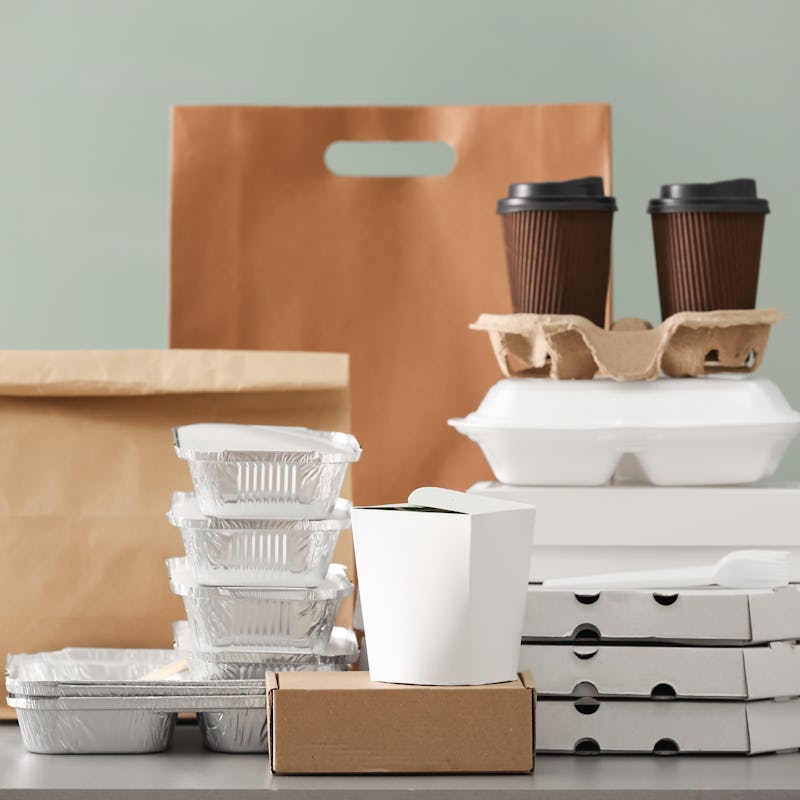 Different packages and carton cups on table against color background. Food delivery service