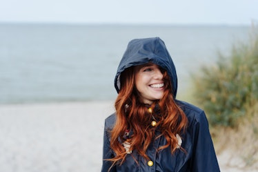 Smiling young red-haired woman in warm blue jacket with a hood on, standing on the beach and looking...