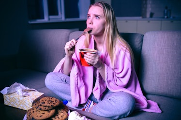 Young woman watching movie at night. Concentrated model eating ice cream with spoon. Sitting on sofa...