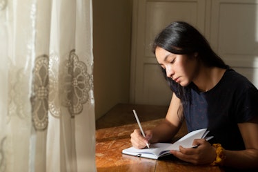 Young Hispanic woman writing in a notebook, serious girl writing in her journal in her room at sunse...
