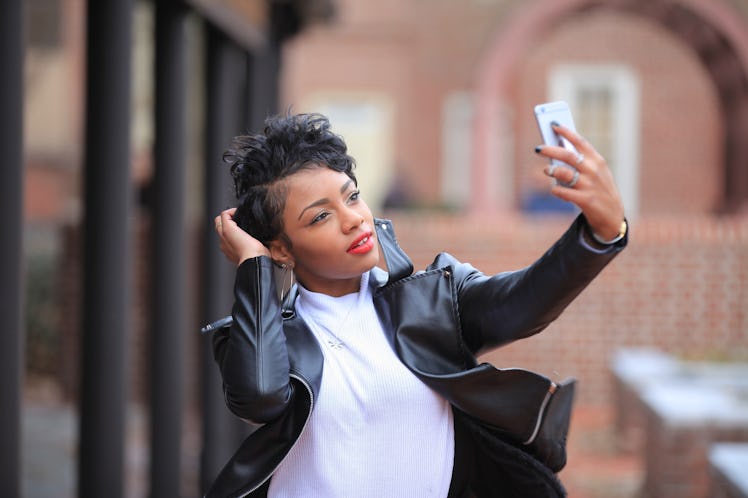 brunette African American woman taking the selfie and smiling with hands near her ears while standin...