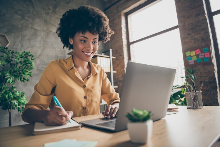 Photo of cheerful joyful mixed-race woman in yellow shirt smiling toothily writing down notes holdin...