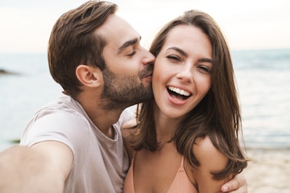 Image of young happy man kissing and hugging beautiful woman while taking selfie photo on sunny beac...