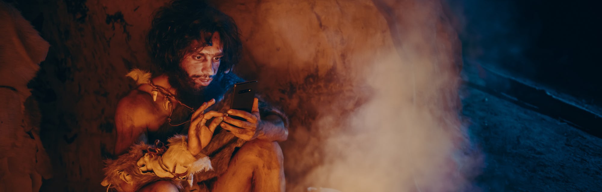 Tribe of Prehistoric, Primitive Hunter Gatherer Wearing Animal Skin Uses Smartphone in a Cave at Night. Neanderthal / Homo Sapiens Male Browsing Internet on Mobile Phone, Watches Videos