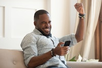 Excited overjoyed black man winner holding smartphone feeling euphoric with mobile online bet bid ga...