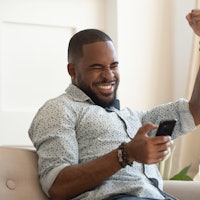 Excited overjoyed black man winner holding smartphone feeling euphoric with mobile online bet bid ga...
