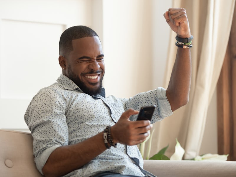 Excited overjoyed black man winner holding smartphone feeling euphoric with mobile online bet bid ga...