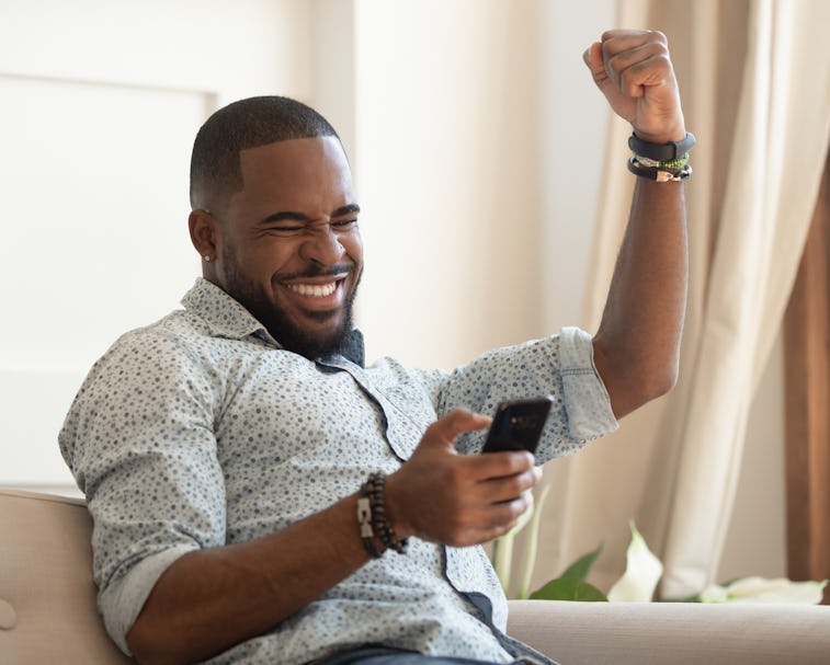 Excited overjoyed black man winner holding smartphone feeling euphoric with mobile online bet bid ga...