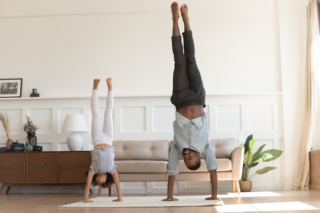 Cute active african kid girl copy imitate father doing gymnastic handstand exercise in living room, ...