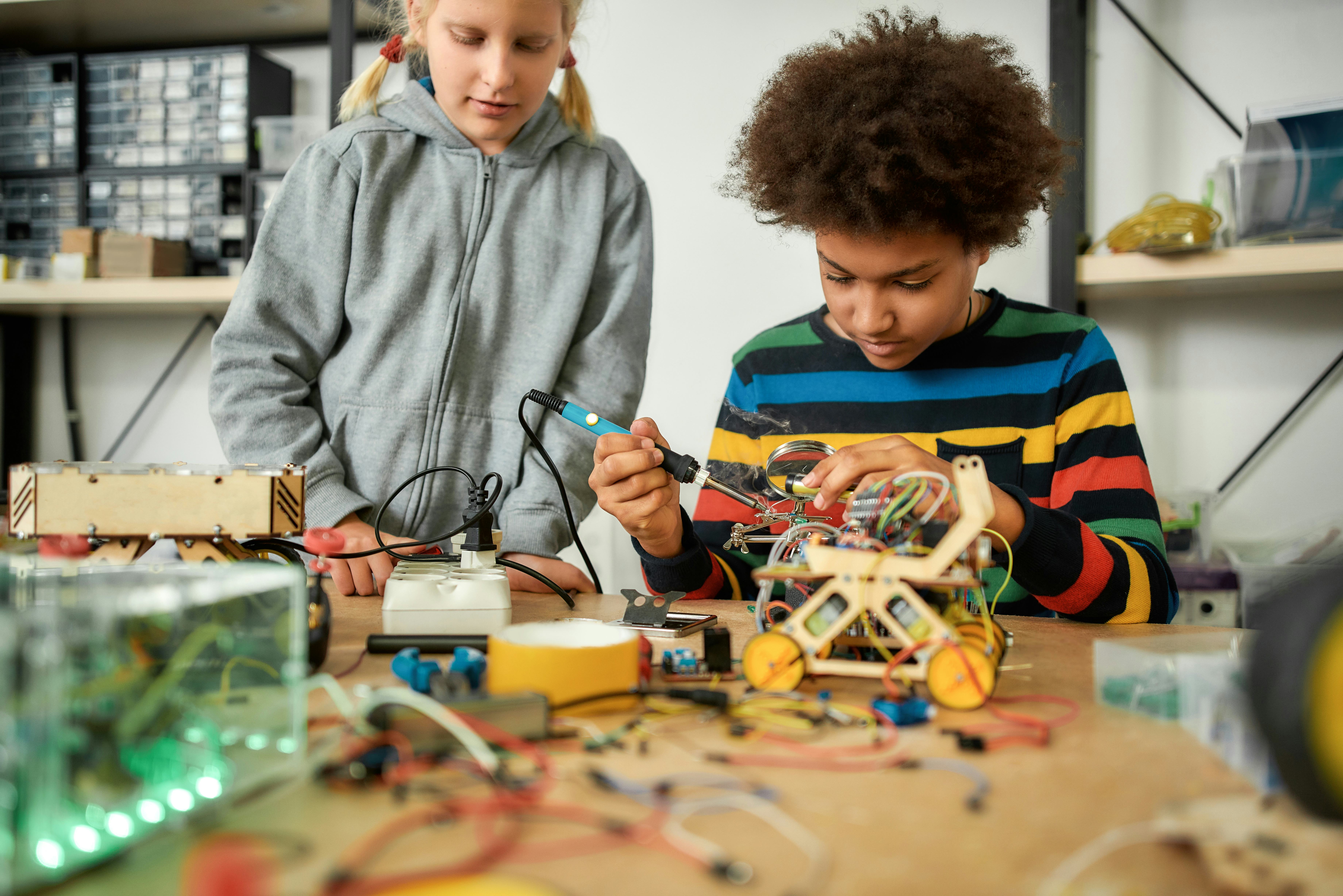Young engineer using soldering iron to join chips and wires. His friend looking at the process. Robo...