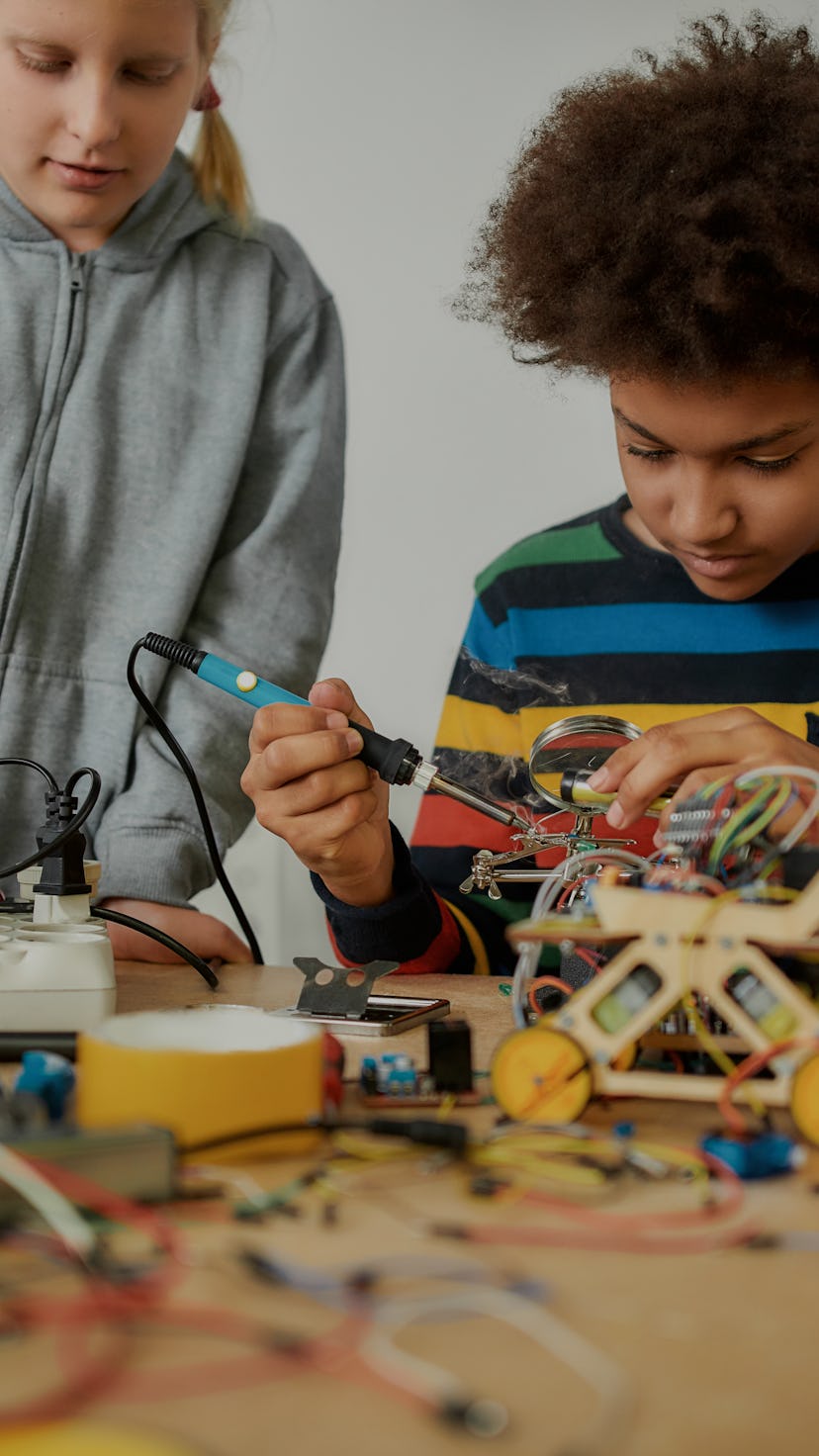 Young engineer using soldering iron to join chips and wires. His friend looking at the process. Robo...