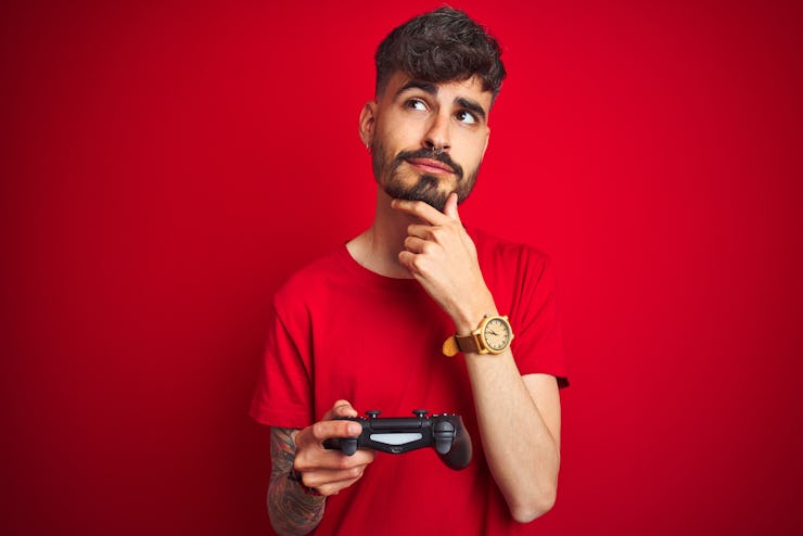 Young gamer man with tattoo playing video game standing over isolated red background serious face th...