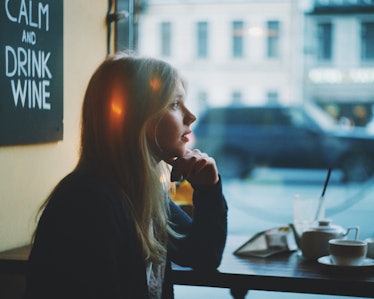 Portrait of lonely blonde woman thinking in a coffee shop and looking through the window at moody ra...