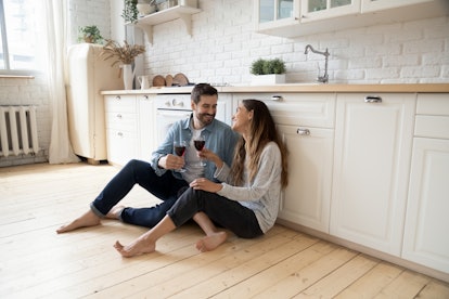 Romantic smiling couple young guy and girl sit on warm kitchen floor talking holding glasses having ...