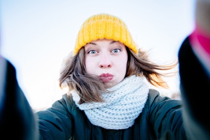 Portrait of young  woman in knitted yellow cap, scarf and mittens taking selfie and heaving fun in w...