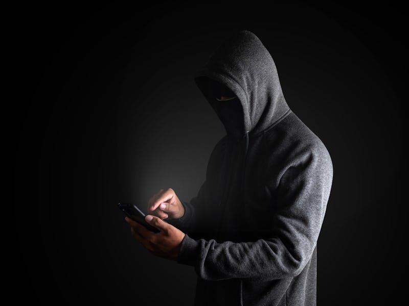 Hacker Using Smartphone. Men in black clothes with hidden face looks at smartphone screen on black b...